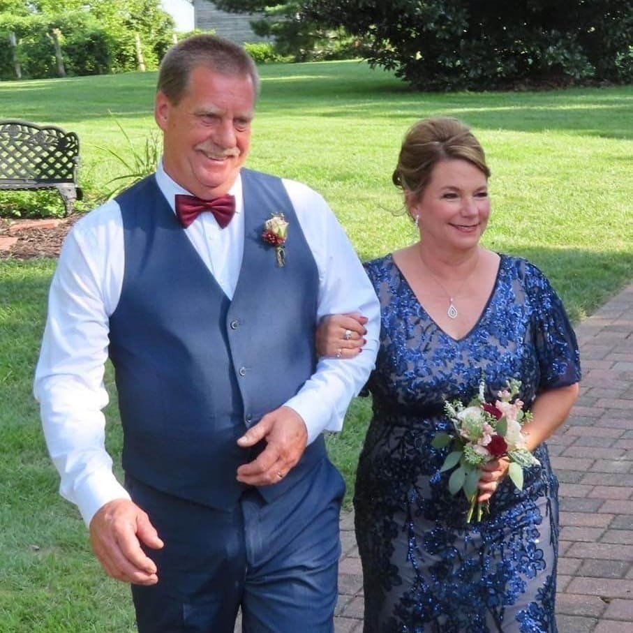 Two people, dressed formally, smiling and walking arm-in-arm on a brick path in a lush garden setting with benches and greenery.